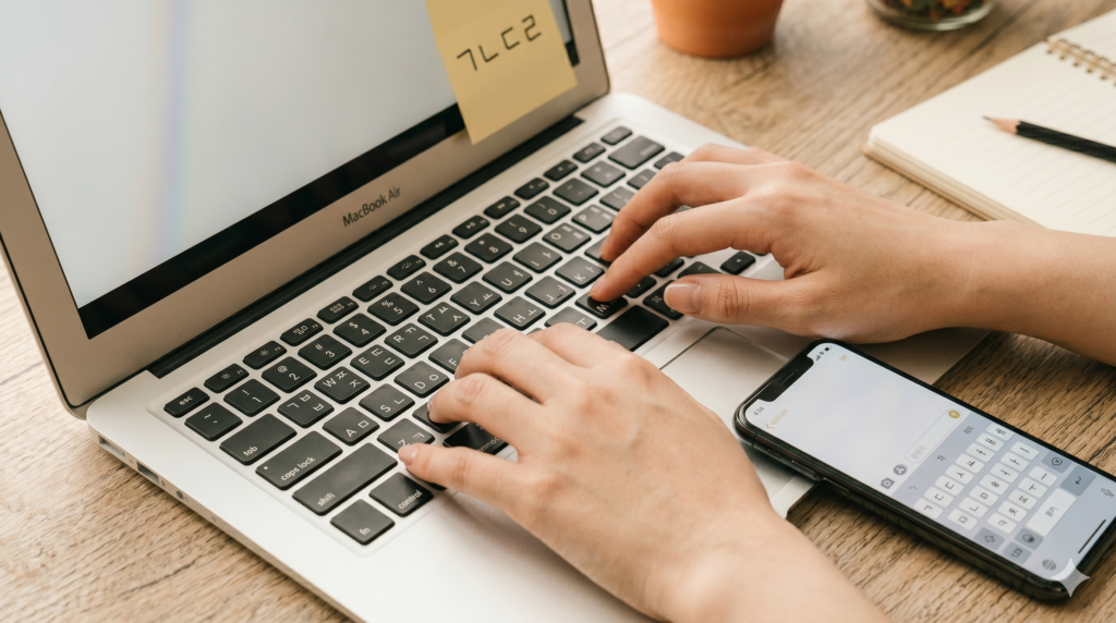 Hands typing on a MacBook with Korean Hangul keyboard, phone showing Korean keyboard beside it on wooden desk