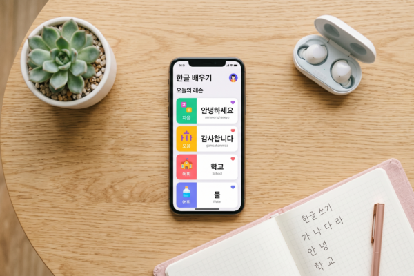 Smartphone displaying a colorful Korean language learning app with Hangul lessons next to a pink notebook with Korean handwriting practice and wireless earbuds on a wooden desk